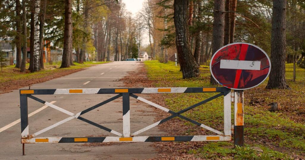 Černo-bílá dopravní závora se žlutými reflexními pruhy uzavírá asfaltovou cestu v lese, vedle ní pokřivená kulatá značka zákaz vjezdu – symbol bariér v digitálním prostředí.